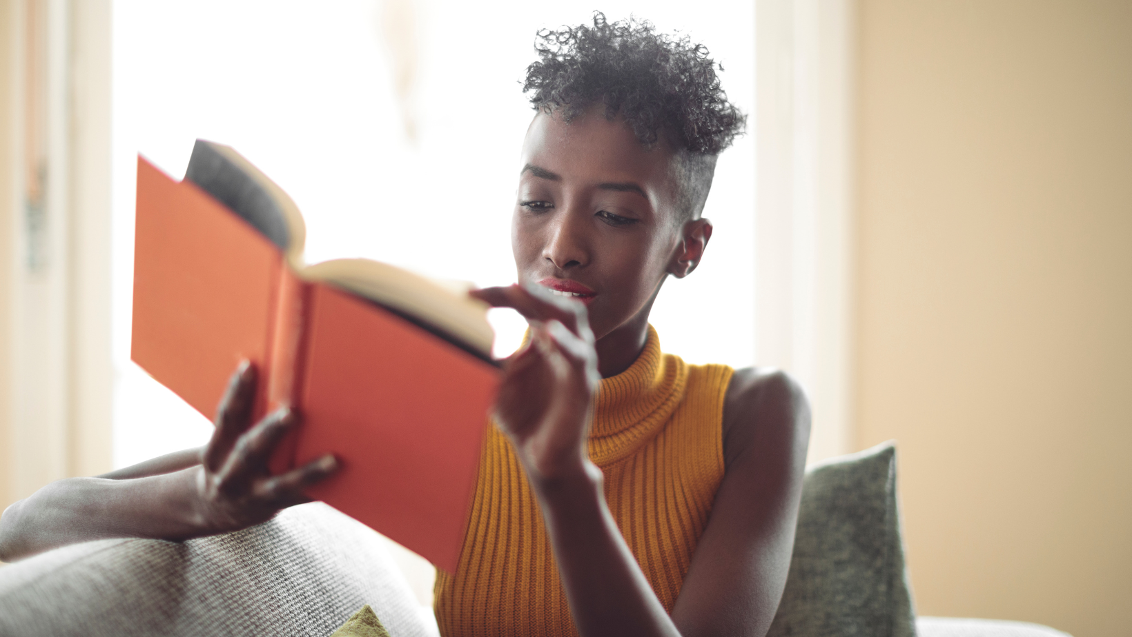 Young Black woman reading an orange book on a couch