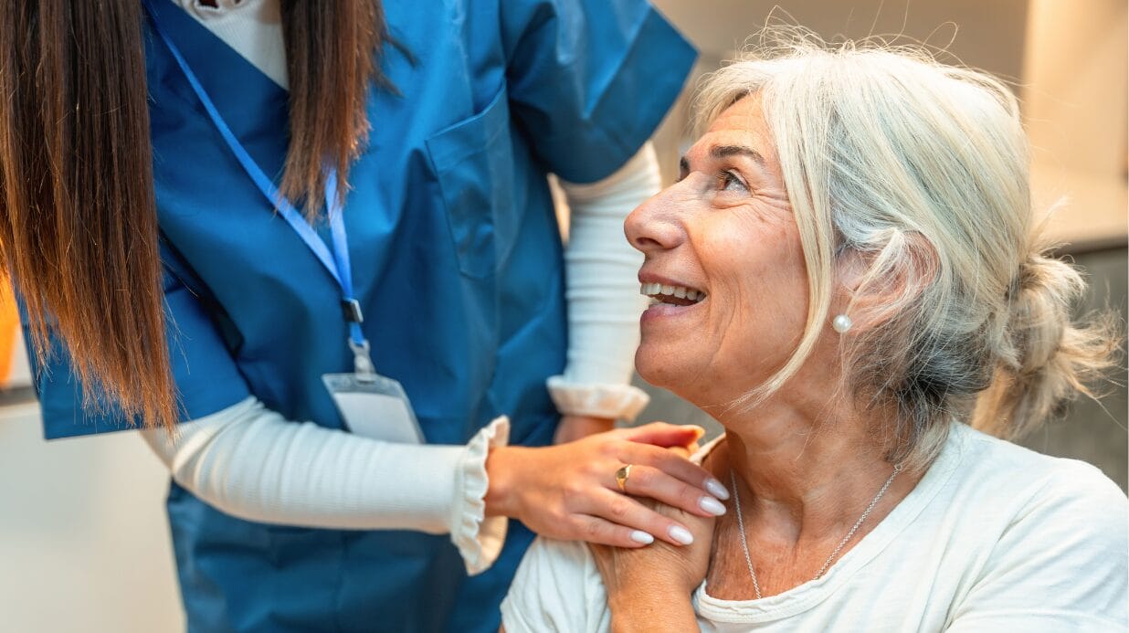 Caregiver with her hand on the shoulder of a senior woman, who is smiling up at the caregiver