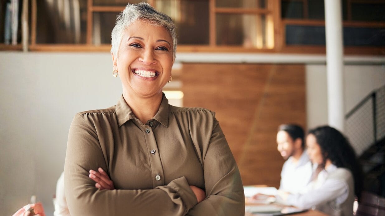 Blog Covers (15) Confident female entrepreneur smiling at the camera with a team meeting in the background