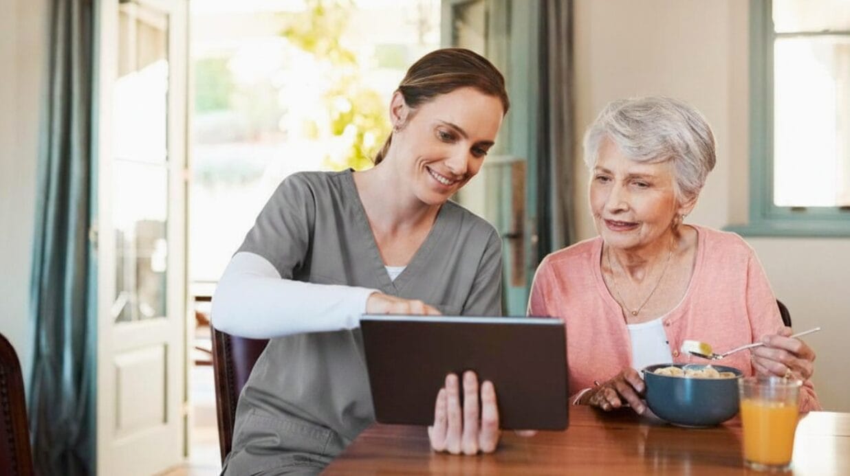 Senior caregiver helping a senior with a tablet while she eats breakfast